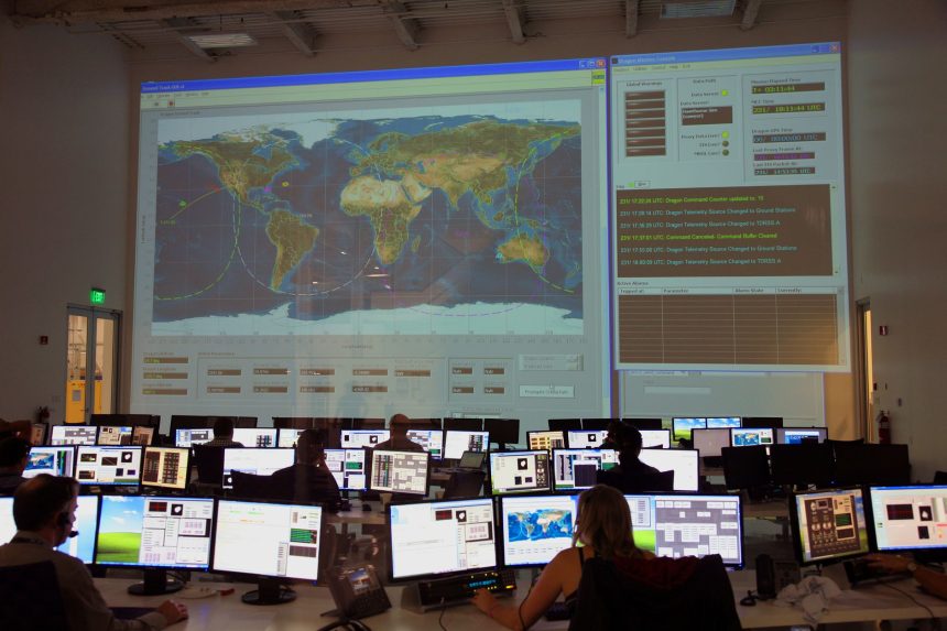 Equipe vista de costas analisa dados em computadores diante de um grande mapa digital em uma sala de controle.